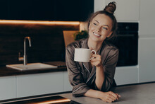 Happy Smiling Young Woman In Brown Satin Pajama Enjoying Aromatic Morning Coffee In Kitchen