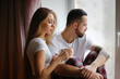 © natalialeb - Portrait of young man and woman drink coffee from mugs hugging and talking on window sill near window. Copy space.Cozy good Christmas morning.