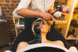 © PoppyPix - Beautician putting a green face mask on her blonde female client laying on a bed. The mask on half of client's face. High quality photo