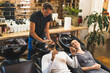 © PoppyPix - Happy indian male client having his hair washed with shampoo by hairdresser next to blonde female customer in a professional hair salon. High quality photo