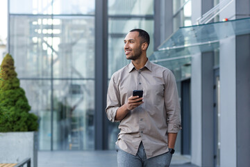 Smiling hispanic man walking down street near modern office building, freelancer businessman looking away holding mobile phone.