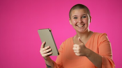 Portrait of happy smiling gender fluid non binary woman promoter 20s shopping online on mobile tablet computer giving thumbs up isolated on pink background studio with copy space