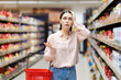High price and inflation. Portrait of young shocked caucasian woman holds cart and takes hand to head. Showcase at background. Person forget to buy something. Concept of shopping in supermarket