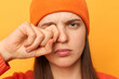 Portrait of unhappy tired exhausted woman wearing orange sweater and beanie hat, rubbing her eyes, frowning face, feels pain or fatigue, standing isolated over yellow background.