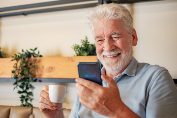 Handsome senior man sitting inside a cafeteria enjoying an espresso coffee cup while using mobile phone - caucasian elderly bearded man looking at smartphone