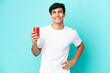 © luismolinero - Young Argentinian man holding a refreshment isolated on blue background posing with arms at hip and smiling