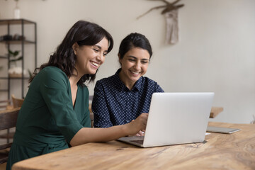 Canvas Print - Two cheerful diverse office workers women cooperating on project, sitting at work desk with laptop, looking at screen, smiling, laughing. Female mentor teaching Indian student, pointing at monitor