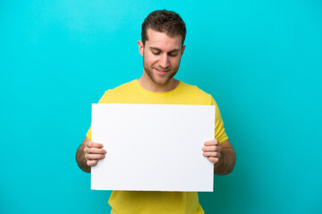 Wall Mural - Young caucasian man isolated on blue background holding an empty placard and looking it