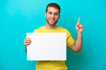 Wall Mural - Young caucasian man isolated on blue background holding an empty placard and pointing up
