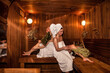 Two women resting leisure in wooden sauna with hot steam. Russian bathhouse. Young females with bath besoms relaxing on bench in spa complex. Wellness, self care, healthy concept. Copy text space