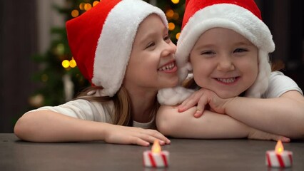 Wall Mural - Happy girls in red santa hat laugh on background of Christmas tree at home. Little children spend New Years Christmas holiday time.