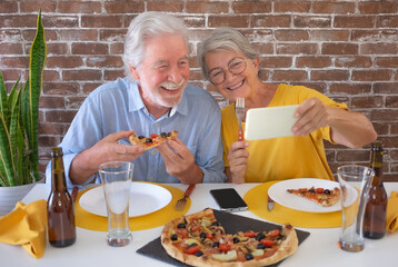 Wall Mural - Happy senior couple having fun together eating a pizza while the woman gets a selfie with phone. Sitting at home table with beer and pizza. Celebration holiday in company