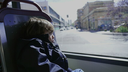 Sad child inside bus public transportation. One depressed little boy staring out window seeing city pass by