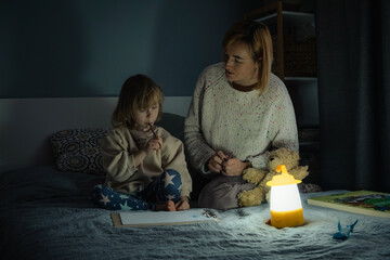 Mother and little daughter studying and drawing in a complete darkness during electricity outage. Little girl uses camping lantern to do her homework during blackout. Energy crisis concept