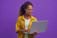 Young Beautiful Happy African American Woman Holding Laptop Visiting Internet Sites Of Shops And Doing Online Shopping During Sales And Discounts Dressed In Casual Clothes Stands In Lilac Studio