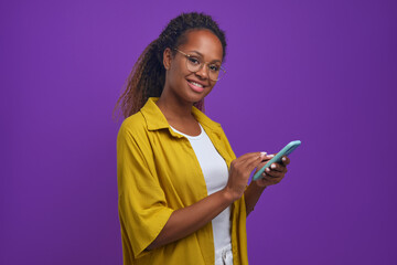 Wall Mural - Young cheerful beautiful African American woman holding phone and looking at camera smiling runs fingers over display of smartphone making purchases via online application stands in lilac studio