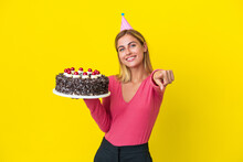 Blonde Uruguayan Girl Holding Birthday Cake Isolated On Yellow Background Pointing Front With Happy Expression