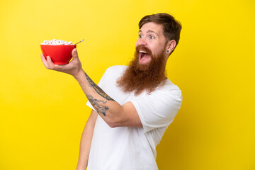 Wall Mural - Redhead man with beard eating a bowl of cereals isolated on yellow background with surprise and shocked facial expression