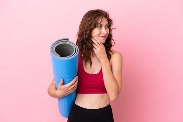 Wall Mural - Young sport woman going to yoga classes while holding a mat isolated on pink background looking up while smiling