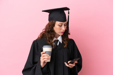 Wall Mural - Young university graduate woman isolated on pink background holding coffee to take away and a mobile