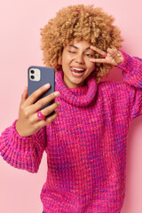 Wall Mural - studio shot of good looking woman sticks out tongue makes peace gesture over eye makes selfie via sm