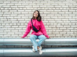 © Raisa Kanareva - Fashion Asian young woman in a pink sweatshirt and jeans sits against a brick wall background, has fun.