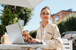 © Sergio - Smiling cute young woman working on laptop while sitting in street cafe on sunny day and looking away. Portrait of business woman freelancer sitting at table outdoors, remote work
