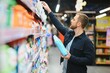 © Serhii - Young man in the supermarket in the household chemicals department. Large selection of products. A brunette in a glasses and a beard in a beige coat.