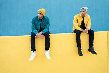 Wall Mural - Funny black twin brothers wearing winter clothes, sitting on yellow and blue wall 