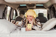 © Renata Hamuda - a laughing woman laying indoor mini camper van using a mobile phone and listening to the music with yellow headphones. Travel vanlife digital nomad people