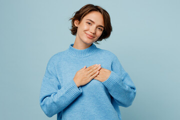 Young greatful happy fun smiling caucasian woman in knitted sweater look camera put folded hands on heart isolated on plain pastel light blue cyan background studio portrait People lifestyle concept