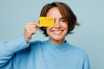 Wall Mural - Close up young smiling happy fun caucasian woman wearing knitted sweater holding in hand cover eye with mock up of credit bank card isolated on plain pastel light blue cyan background studio portrait.