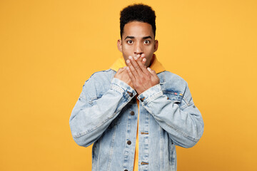 Young scared fearful sad amazed man of African American ethnicity in denim jacket hoody look camera cover mouth with hand isolated on plain yellow background studio portrait People lifestyle concept