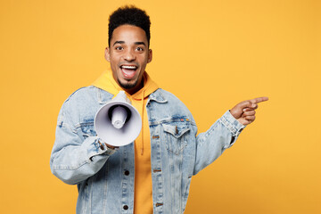 Wall Mural - Young shocked excited man of African American ethnicity wear denim jacket hoody hold in hand megaphone scream announces discounts sale Hurry up point aside isolated on plain yellow background studio.