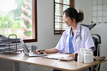 Wall Mural - Attractive Asian female doctor using calculator and laptop, estimating surgery cost for her patient