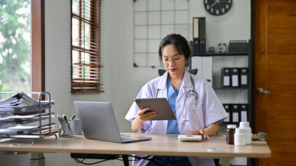 Beautiful Asian female doctor using calculator and digital tablet, working in the office.