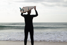 Back View Of Elderly Surfer With Board Before Surfing. Gray-haired Man Holding Board On Head Standing On Sandy Sea Shore Looking At Sea To Go Into Water. Active Rest And Sport For Aged People Concept