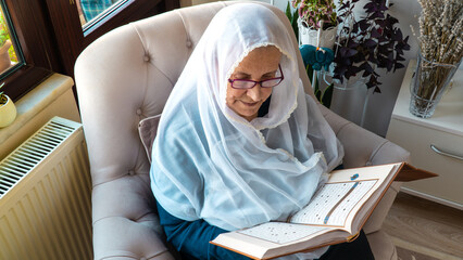 Old Muslim woman reading the Holy Quran at home during Ramadan Month. Old woman wearing white hijab reading the Koran.