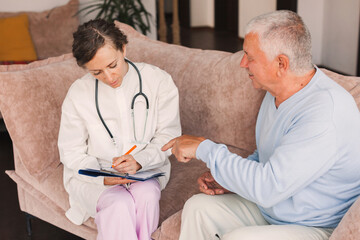 Female professional doctor showing medical test result explaining prescription using clipboard visiting senior elderly old man patient at home sitting on sofa