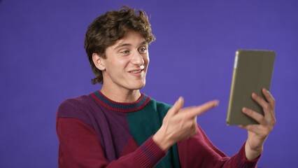 Portrait of happy smiling young guy 20s in sweater having video chat on tablet computer conducting pleasant conversation isolated on purple background studio. 