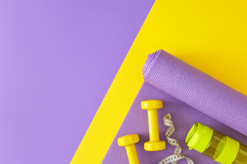 Creative sports concept. Top view composition of dumbbells, exercise mat, tape measure, bottle of water on yellow and violet  background with copy space. Minimal fitness idea.