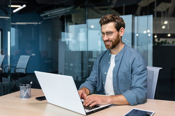 Smiling and cheerful businessman in casual shirt working inside modern office, mature programmer writing code using laptop.