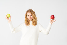 A Young Pretty Curly Blonde Woman Holds A Yellow Green And Red Apple In Her Hands. The Girl Looks Happy And Smiles, She Is Dressed In A White Knitted Suit. Near The Model There Is A Lot Of Space For A
