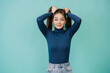 Young adult pretty woman in blue sweater, jeans looks at camera, holding her hair, making two ponytails over turquoise studio backdrop with empty space. Mockup. Cute Swedish girl with healthy hair.