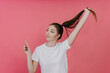 © Iona - Healthy, well-groomed young Italian girl in a white T-shirt, holding her ponytail out of her hair, pointing high with her index finger at an empty place, standing in the studio on a pink background.