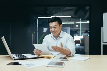 Wall Mural - A young man, an Asian businessman, sits smiling in the office at the table, works with documents, holds papers, contracts, bills in his hands.