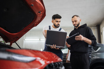 Close up of two young bearded engineer mechanic under car hood checking engine with laptop and tablet using electronic diagnostic equipment to tune sport red vehicle.