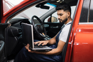 Wall Mural - Bearded male mechanic sitting inside car using laptop, recording automobile engine checks collect detailed information during his work on car workshop. Service maintenance during engine repair.