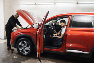 Wall Mural - Young male mechanic using laptop sitting inside red car and his colleague with tablet under hood, recording automobile engine checking collecting detailed information during their work on car workshop