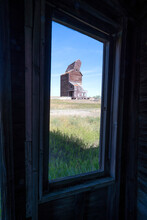 Crumbling Grain Elevator Viewed Through The Window Of An Old Abandoned Building In Rural Saskatchewan; Bents, Saskatchewan, Canada